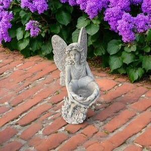 May include: A gray stone angel statue with detailed wings and a floral bowl. The angel is seated on a bed of flowers, set against a backdrop of purple flowers and a brick walkway.