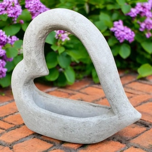 May include: A gray, heart-shaped planter made of concrete. The planter has a curved handle and a shallow basin for holding plants. The background features purple flowers and green foliage, set on a brick surface.