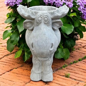 May include: A gray concrete moose-shaped planter with a large opening for plants. The moose has large antlers, a detailed face, and a textured surface. The planter is surrounded by green leaves and purple flowers.