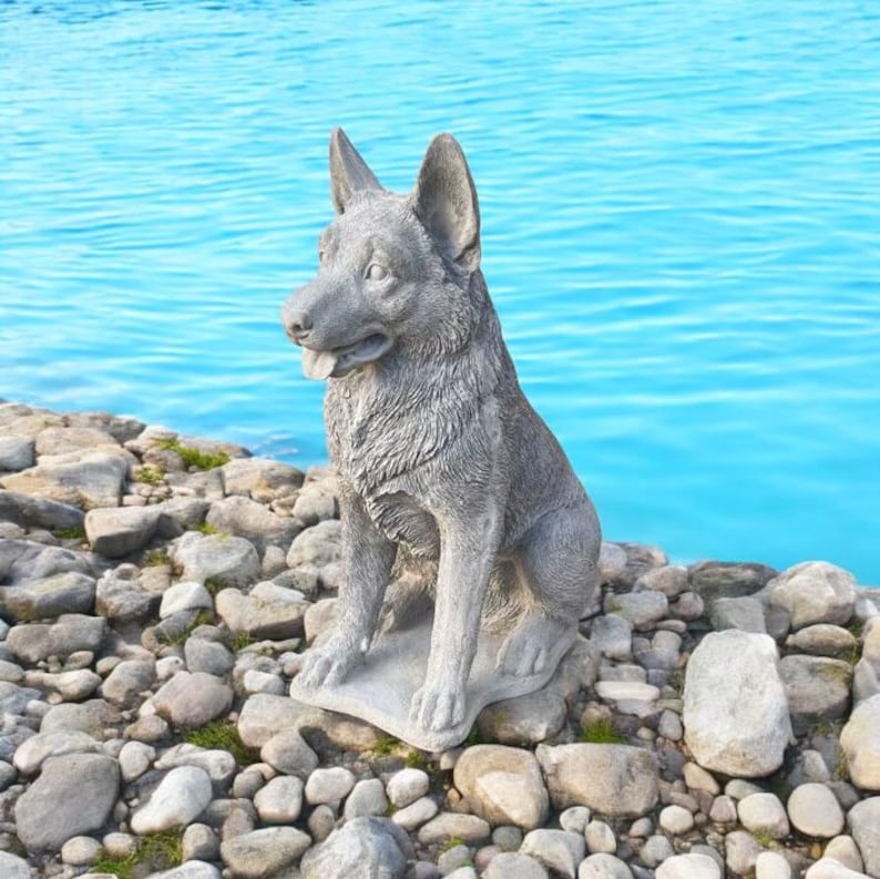 Pu&ograve; includere: Una statua di un pastore tedesco in cemento grigio seduta su un letto di pietre grigie. Il cane guarda a sinistra e ha un aspetto realistico.
