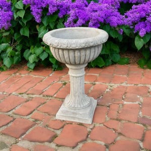 May include: A gray stone planter with a fluted pedestal base and a decorative rim. The planter is set on a brick patio, with a backdrop of vibrant purple flowers and green foliage.