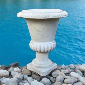 May include: A white stone urn with a decorative rim sits on a bed of grey stones in front of a blue body of water.