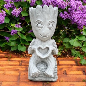 May include: A gray statue of a character with a smiling face and large eyes, holding a heart shape with its hands. The statue is seated and has a textured appearance, resembling wood. The background features vibrant purple flowers and green foliage.