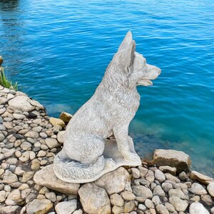Pu&ograve; includere: Una statua di cane in cemento grigio seduta vicino a un specchio d'acqua. Il cane guarda a destra e ha un design di pelliccia testurizzato.