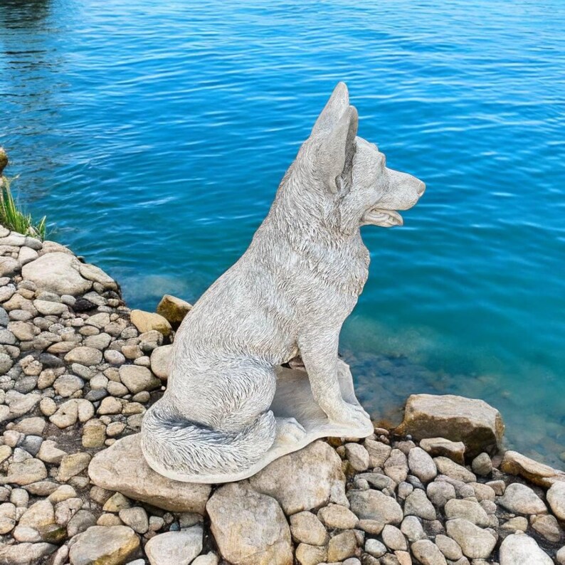 Pu&ograve; includere: Una statua di cane in cemento grigio seduta vicino a un specchio d'acqua. Il cane guarda a destra e ha un design di pelliccia testurizzato.