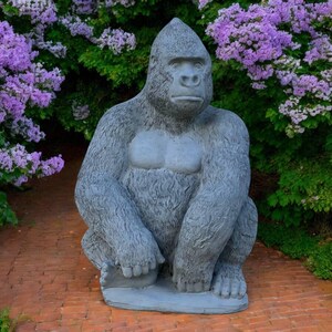 May include: A gray gorilla statue with a seated pose, featuring a detailed fur texture and a lighter chest. The sculpture is placed on a brick surface, surrounded by lush green foliage and vibrant purple flowers, creating a garden decor theme.