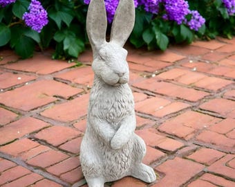 Può includere: Una statua di coniglio in cemento grigio con lunghe orecchie e una superficie strutturata. Il coniglio è in piedi con le zampe incrociate, sullo sfondo di fiori viola e pavimenti in mattoni rossi. Un ornamento da giardino.