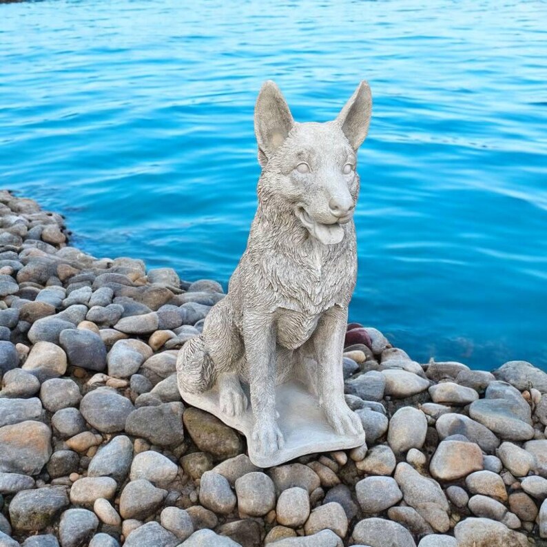 Pu&ograve; includere: Una statua di un pastore tedesco in cemento grigio seduta su un letto di ciottoli grigi. Il cane guarda a destra con la lingua di fuori.