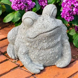 May include: A gray concrete frog statue with textured skin, large eyes, and a wide mouth. The frog is sitting on a brick surface, surrounded by green leaves and vibrant purple flowers. A garden ornament.