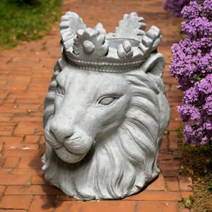 May include: A grey concrete lion head planter, adorned with a crown. The lion's features are detailed, including a mane and facial characteristics. The planter is designed for outdoor use and is positioned on a brick surface. The background features purple flowers.
