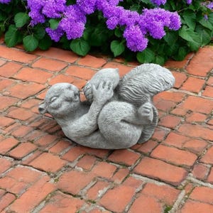 May include: A grey concrete squirrel statue in a playful pose, holding a nut. The squirrel is lying on its back on a brick surface, with purple flowers and green foliage in the background. A charming garden ornament.