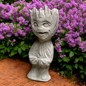 May include: A grey, sculpted planter in the shape of a character with a smiling face and textured details. The planter is standing on a brick surface, with a backdrop of vibrant purple flowers and green foliage.