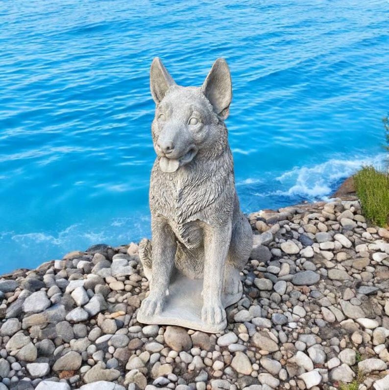 Pu&ograve; includere: Una statua di un pastore tedesco in cemento grigio, seduto con la lingua di fuori. La statua &egrave; posizionata su un letto di piccole pietre grigie.