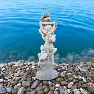 May include: A gray stone sculpture of three frogs stacked on top of each other. The top frog is wearing a hat and holding a sign that says "Welcome". The sculpture is sitting on a bed of gray rocks.