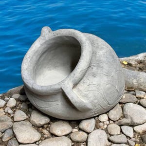 Peut inclure: Un pot en béton gris en forme de pot avec une large ouverture. Le pot est posé sur un lit de pierres grises.