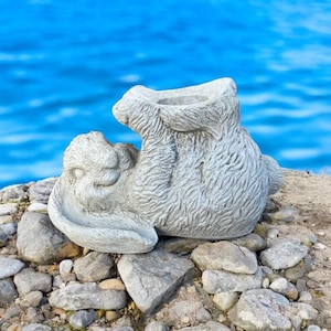 May include: A gray concrete rabbit planter with a textured surface. The planter is lying on its side with its head resting on a pile of small gray rocks.