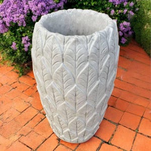 Peut inclure: Un grand pot de fleurs en béton gris avec un motif de feuilles texturées. Le pot de jardin extérieur est de forme ovale et repose sur un patio en briques, avec des fleurs violettes en arrière-plan.