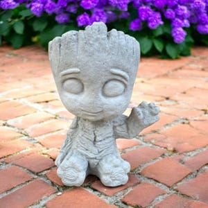 May include: A gray concrete planter shaped like a cartoon character with large eyes and a smiling expression. The planter has a textured surface and is sitting on a brick patio with purple flowers in the background.