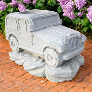 May include: A gray concrete planter shaped like a vintage off-road vehicle. The planter has a detailed design of a car with a square body, wheels, and a front grill. It sits on a rocky base, perfect for outdoor decor. The background features purple flowers.