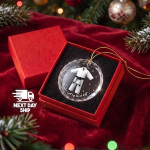 May include: A round, clear, faceted glass ornament featuring a white karate gi with a black belt, printed with the text 'The year I got my black belt' and '2026'. The ornament includes a gold hanging cord and is presented in a red gift box.