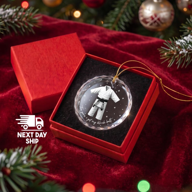 May include: A round, clear, faceted glass ornament featuring a white karate gi with a black belt, printed with the text 'The year I got my black belt' and '2026'. The ornament includes a gold hanging cord and is presented in a red gift box.