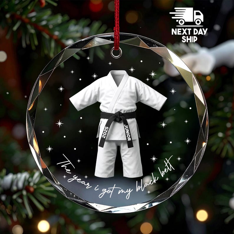 May include: A round, faceted glass ornament featuring a white martial arts uniform with a black belt. The belt is printed with the text 2025 and JUNIOR. Below the uniform, the text reads The year i got my black bett, surrounded by white stars.