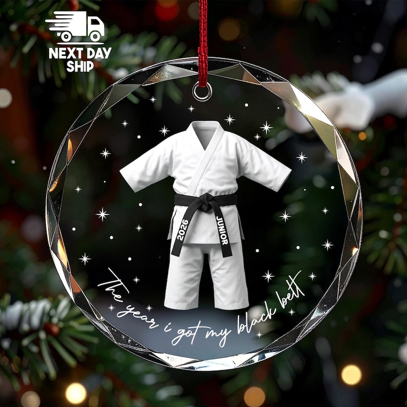 May include: A circular, faceted glass ornament featuring a white martial arts uniform with a black belt. The belt is printed with the text 2026 and JUNIOR. Below the uniform, the text reads The year i got my black bett, surrounded by stars.
