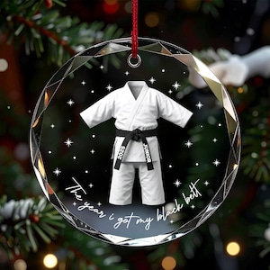 May include: A circular, faceted glass ornament featuring a white martial arts uniform with a black belt. The belt is printed with the text 2025 and JUNIOR. Below the uniform, the text reads The year i got my black belt, surrounded by stars.