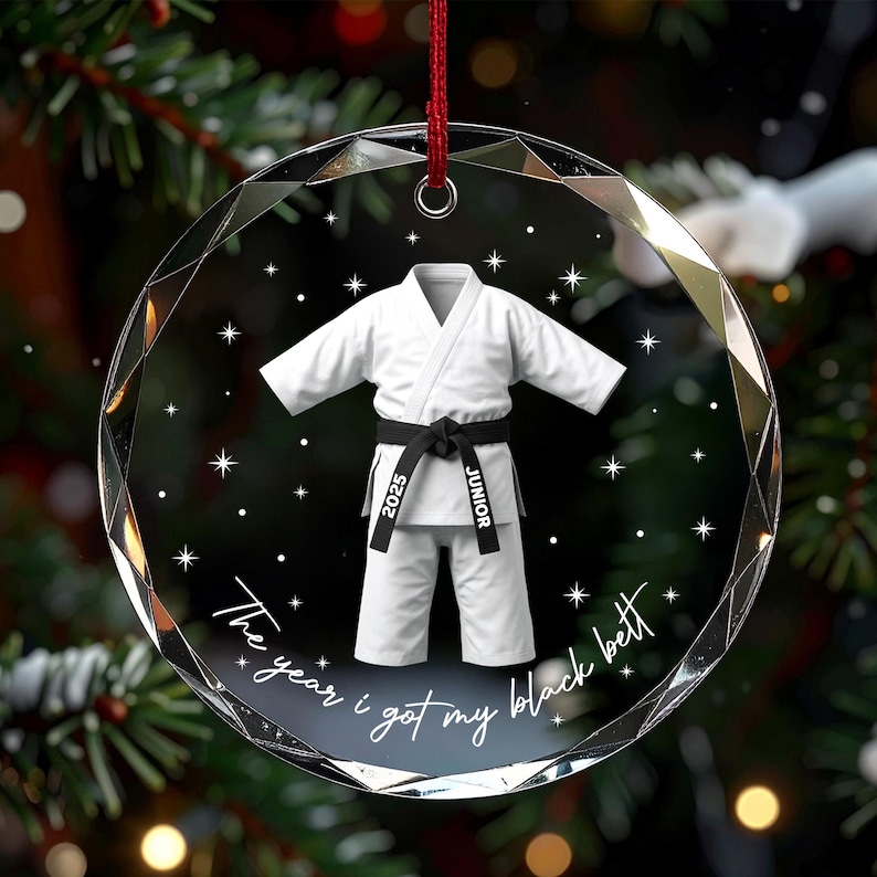 May include: A circular, faceted glass ornament featuring a white martial arts uniform with a black belt. The belt is printed with the text 2025 and JUNIOR. Below the uniform, the text reads The year i got my black belt, surrounded by stars.