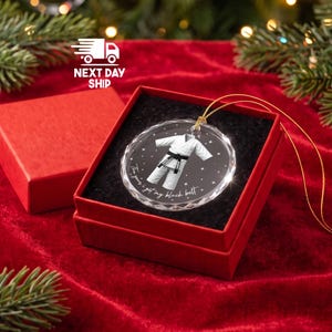 May include: A round, clear glass ornament featuring a white martial arts uniform with a black belt. The design includes small white stars and the text, The year I got my black belt. The ornament is displayed inside a square red gift box with black lining.