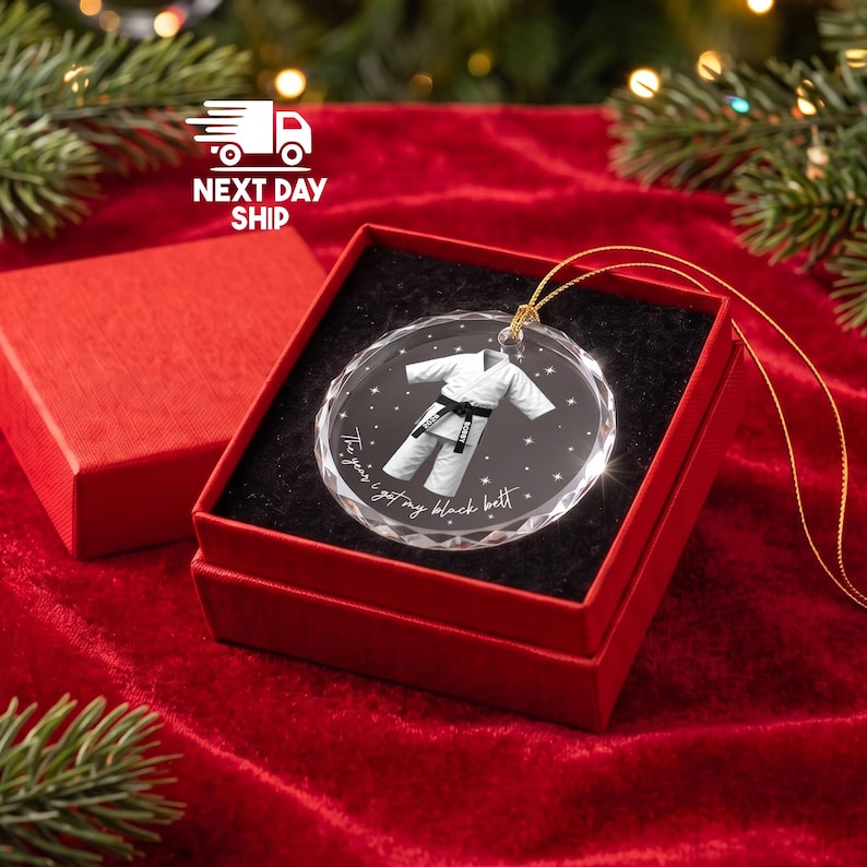 May include: A round, clear glass ornament featuring a white martial arts uniform with a black belt. The design includes small white stars and the text, The year I got my black belt. The ornament is displayed inside a square red gift box with black lining.