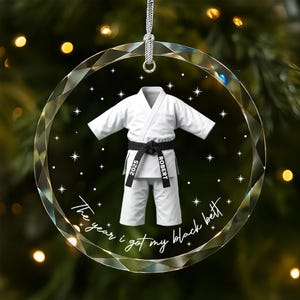 May include: A circular, faceted glass ornament featuring a white martial arts uniform with a black belt. The belt is printed with the text 2025 and ROBERT. Below the uniform, the text reads The year i got my black belt with stars.