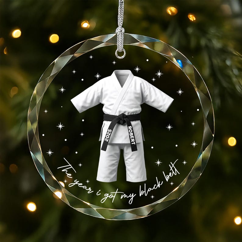 May include: A circular, faceted glass ornament featuring a white martial arts uniform with a black belt. The belt is printed with the text 2025 and ROBERT. Below the uniform, the text reads The year i got my black belt with stars.