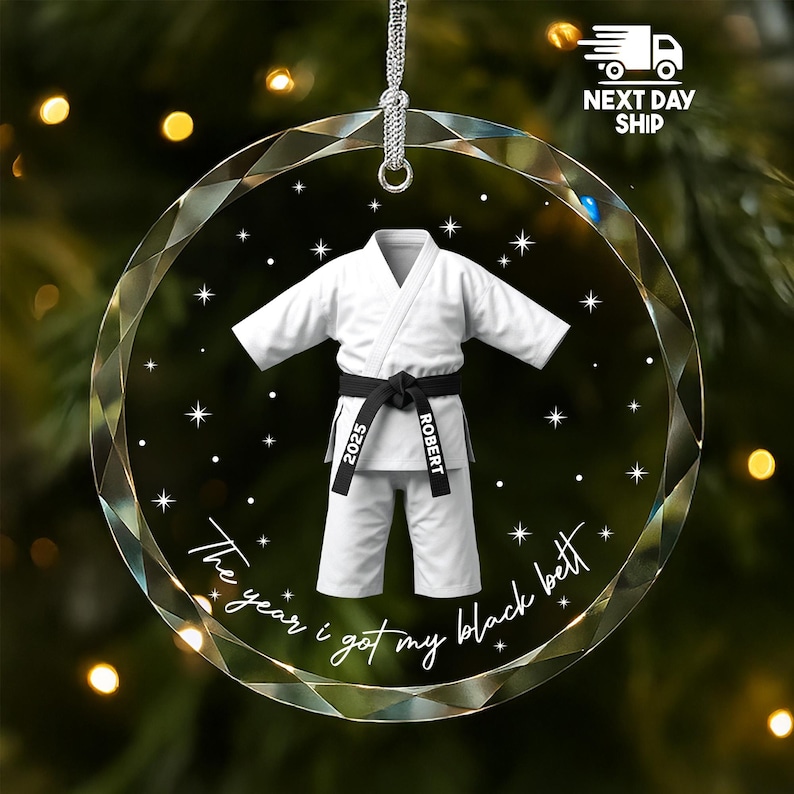 May include: A circular, faceted glass ornament featuring a white martial arts uniform with a black belt. The belt is printed with the text 2025 and ROBERT. Below the uniform, the text reads The year i got my black belt, surrounded by stars.