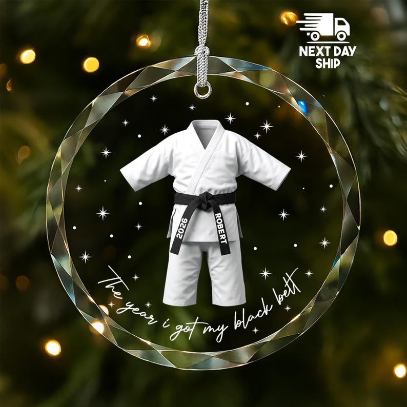 May include: A circular clear acrylic ornament featuring a white martial arts uniform with a black belt. The belt is printed with the text 2026 and ROBERT. Below the uniform, the text reads The year i got my black belt, surrounded by white stars.