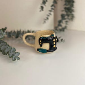 以下が含まれることがあります： 黒とティール色のミシン模様のベージュ色のセラミックマグカップ。マグカップは湾曲したハンドルと金色のアクセントが付いています。ミシンのデザインには、金色のボタンとティール色のディテールが含まれています。ユニークで装飾的なアイテムです。
