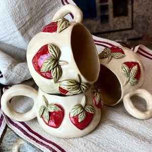May include: Three ceramic mugs featuring strawberry designs. The mugs are white with red and green details. The mugs are stacked on top of each other.