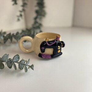 May include: A cream ceramic mug featuring a black sewing machine design with purple and gold detailing. The mug has a rounded handle and a small thread spool detail. The background is a soft white.