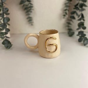May include: A cream-colored ceramic mug with a gold-colored letter "G" on the front. The mug has a textured surface and a rounded handle. The rim of the mug is also trimmed in gold. The mug is a decorative item.