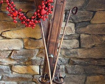 May include: A pair of vintage wooden skis with brown leather straps and two wooden ski poles with black tips. The skis are leaning against a stone wall with a red heart-shaped wreath hanging on the wall.