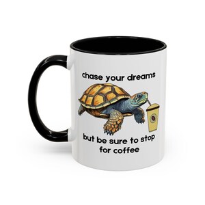 Könnte beinhalten: Weiße Keramik-Kaffeetasse mit schwarzem Innenraum und Griff. Die Tasse zeigt eine Zeichentrickschildkröte mit einer Kaffeetasse und dem Text "chase your dreams but be sure to stop for coffee."