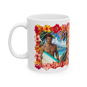 May include: White ceramic mug with a tropical design. The design features a man with a surfboard, flowers, palm trees, and the words "Beach Bum Out Getting Lei'd". The mug has a curved handle.