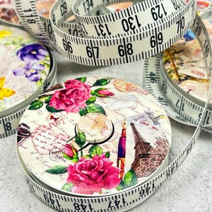 Peut inclure: Un ruban à mesurer blanc avec des marques noires est enroulé autour d'un récipient rond décoratif. Le récipient est orné de roses roses, de papillons et d'illustrations de style vintage. Le ruban mesure jusqu'à 378 cm.
