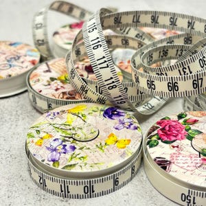 Könnte beinhalten: Mehrere dekorative Maßbandetuis mit Blumen- und Schmetterlingsmotiven. Die weißen Maßbänder sind ausgezogen und zeigen Zentimeter- und Zollmarkierungen. Die Etuis zeigen rosa, lila und gelbe Blumen und Schmetterlinge.