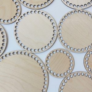 May include: A collection of light-colored wooden basket bases with evenly spaced holes around the perimeter. The circular bases vary in size and are arranged against a white background. The wood grain is visible on the surface of the bases.