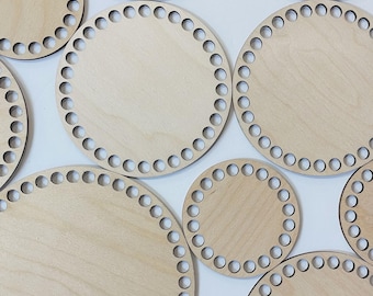 May include: A collection of light-colored wooden basket bases with evenly spaced holes around the perimeter. The circular bases vary in size and are arranged against a white background. The wood grain is visible on the surface of the bases.