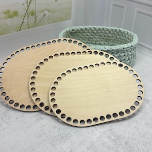 Op de afbeelding: Drie ovale, onafgewerkte houten mandbodems met voorgeboorde gaten voor haakprojecten. Op de achtergrond staat een lichtgroene gebreide mand. De houten bodems zijn lichtbruin.