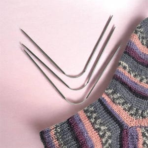 May include: Three silver sock knitting needles are arranged in a V-shape on a light pink surface. A portion of a gray, pink, and purple patterned sock is visible in the lower right corner, suggesting a knitting project.