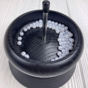 May include: A black wooden bead organizer with a rotating base and a central spindle. White beads are arranged around the spindle in the bowl.