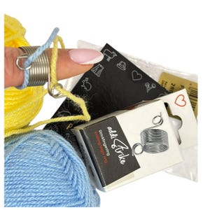 Puede incluir: Primer plano de suministros de tejido, incluyendo lana azul y amarilla, un anillo de tejido y una pequeña caja con la etiqueta "addiTrike Knitting finger ring". También se ve una etiqueta negra con ilustraciones blancas.